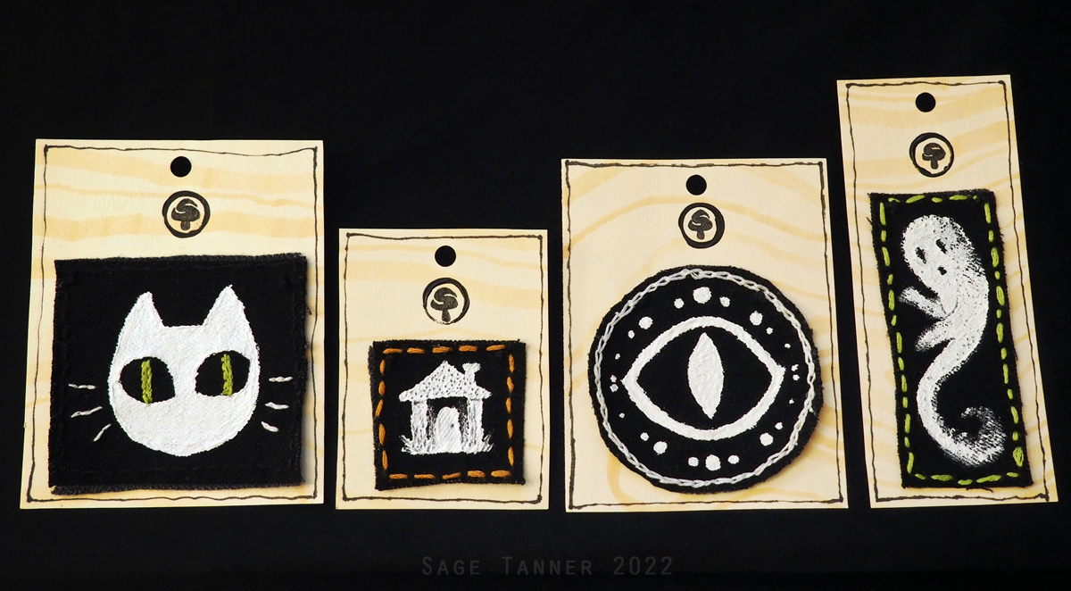 A series of black denim patches with white paint and colored embroidery. A cat, a house, an eye, and a ghost.