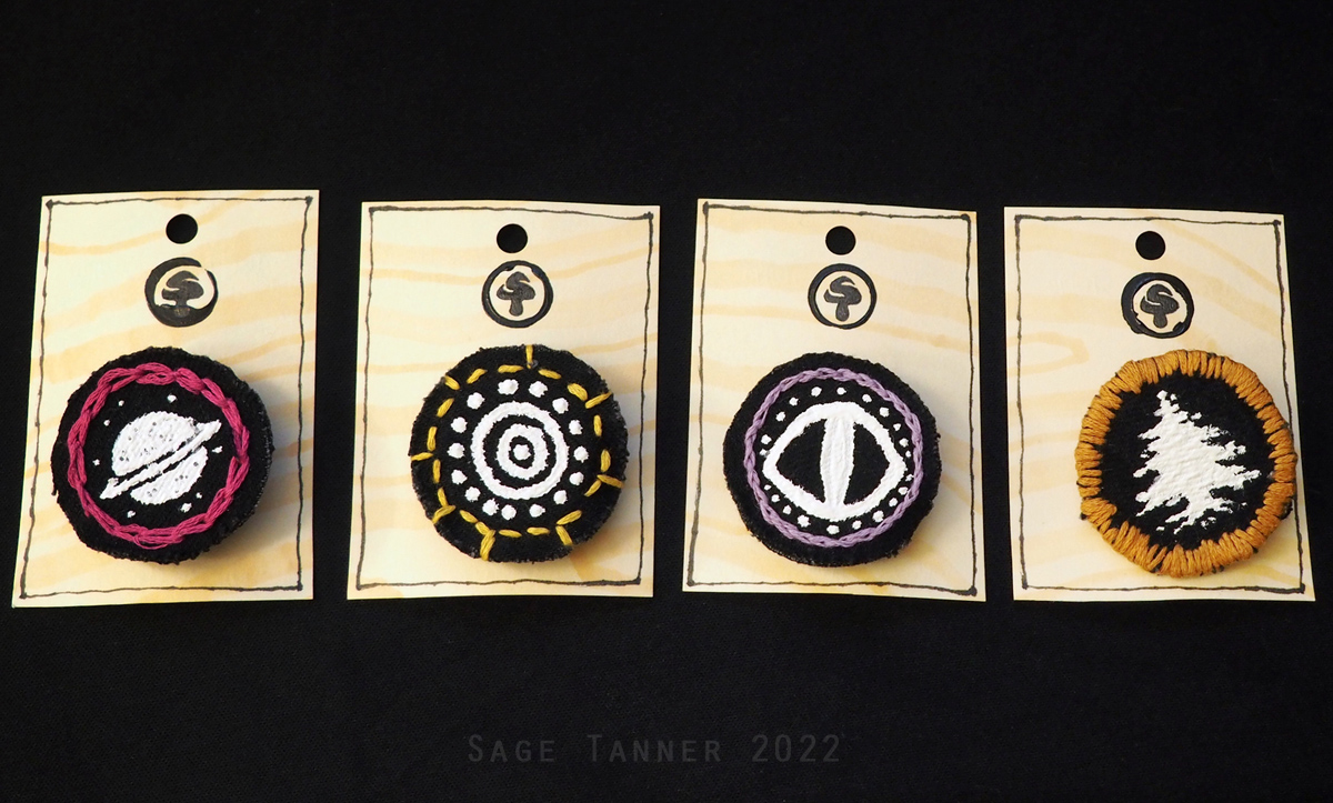 A series of fabric pins with black and white designs. A planet, the sun, an eye, and a tree.